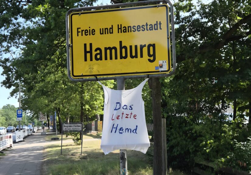 Ein Weisses T-Shirt mit der Aufschrift das letzte Hemd unter dem Ortseingangsschld Hambugr