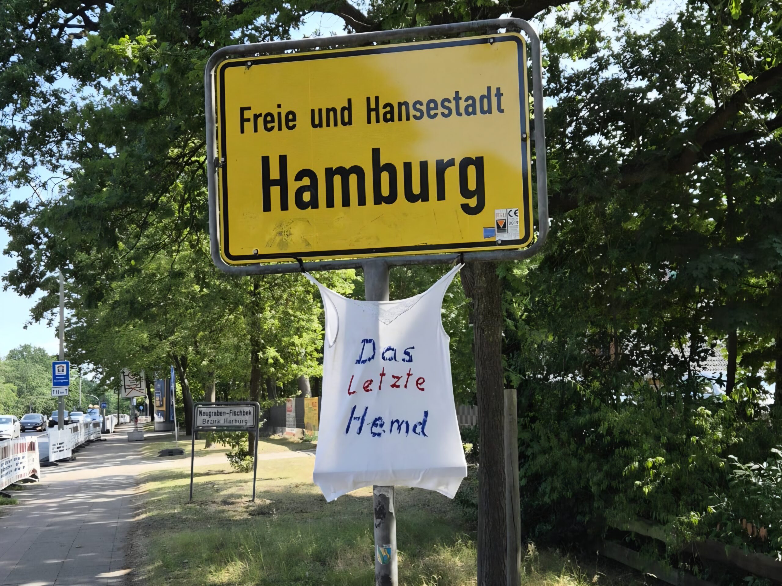 Ein Weisses T-Shirt mit der Aufschrift das letzte Hemd unter dem Ortseingangsschld Hambugr